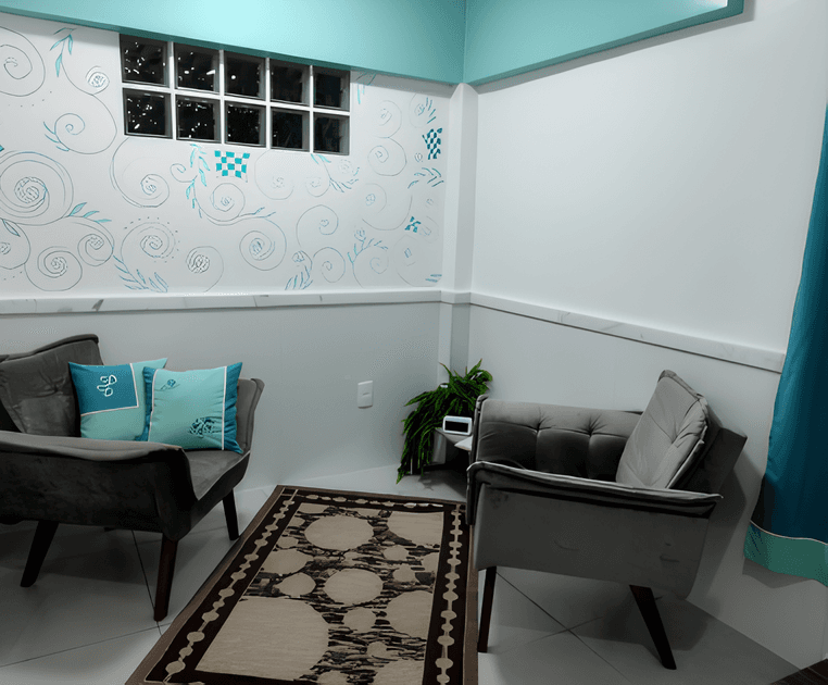 Uma sala pequena e aconchegante com duas cadeiras estofadas na cor cinza, uma delas com almofadas de tecido estampado com detalhes verdes e brancos. A sala tem uma parede branca com um desenho de arabescos verde-claro e um tapete marrom com desenhos circulares.