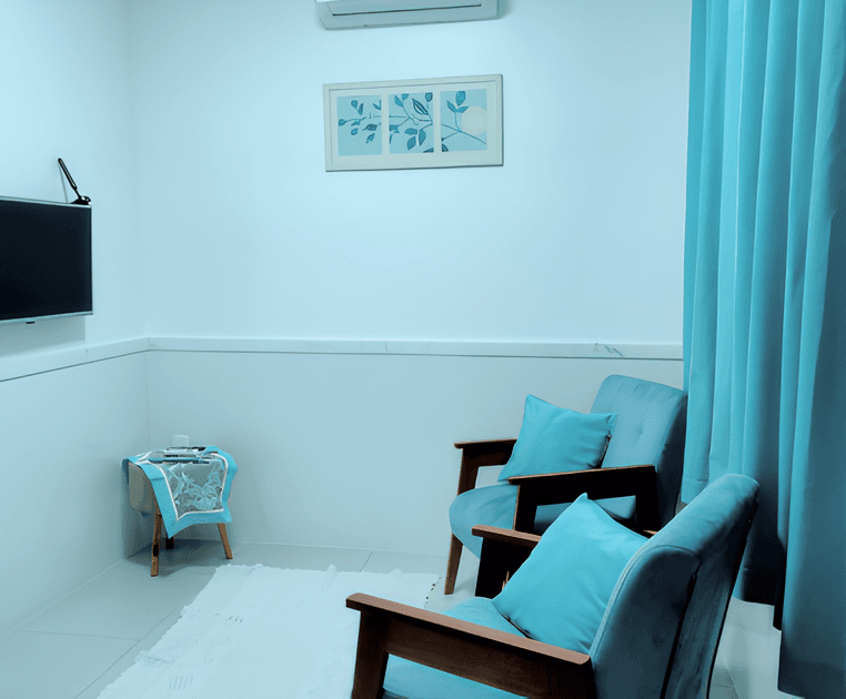 Uma sala pequena e aconchegante com duas cadeiras estofadas azuis, uma mesa de centro com um livro e uma xícara de café, um tapete branco e uma televisão pendurada na parede branca.