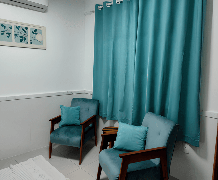 Uma sala pequena com duas cadeiras estofadas verdes com almofadas verdes, uma cortina verde atrás das cadeiras, um tapete branco, uma mesa de centro com um livro e um vaso com flores. A parede branca possui um quadro com um desenho de ramos e uma folha.