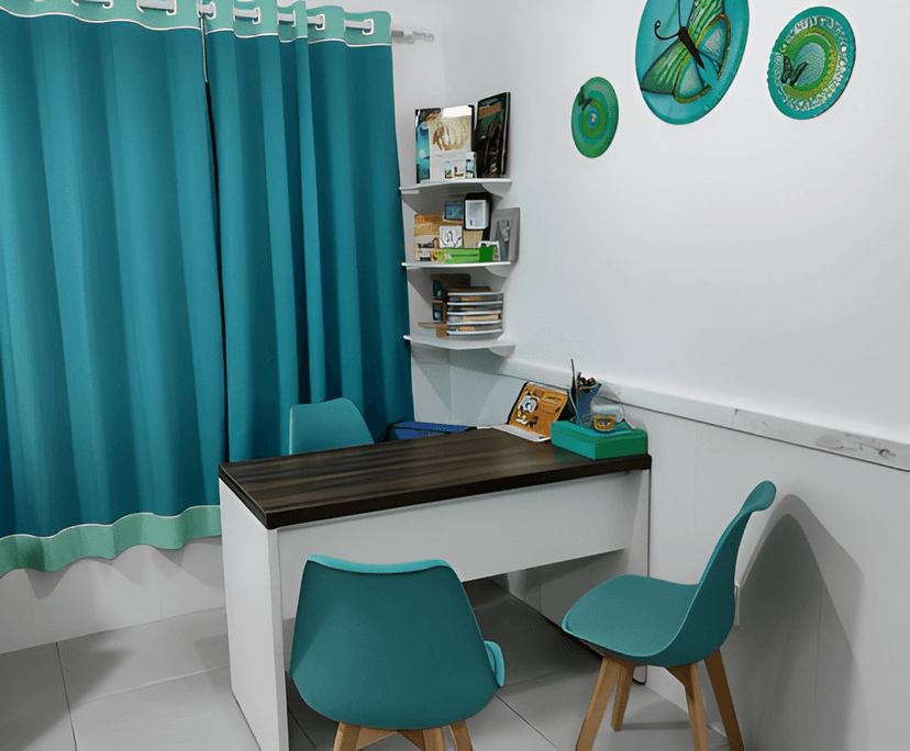 Uma sala com uma mesa de madeira escura e duas cadeiras verdes, com cortinas verdes na parede atrás da mesa. Há uma prateleira de madeira na parede à direita com diversos objetos coloridos, e três círculos com borboletas azuis em um círculo verde na parede à esquerda.