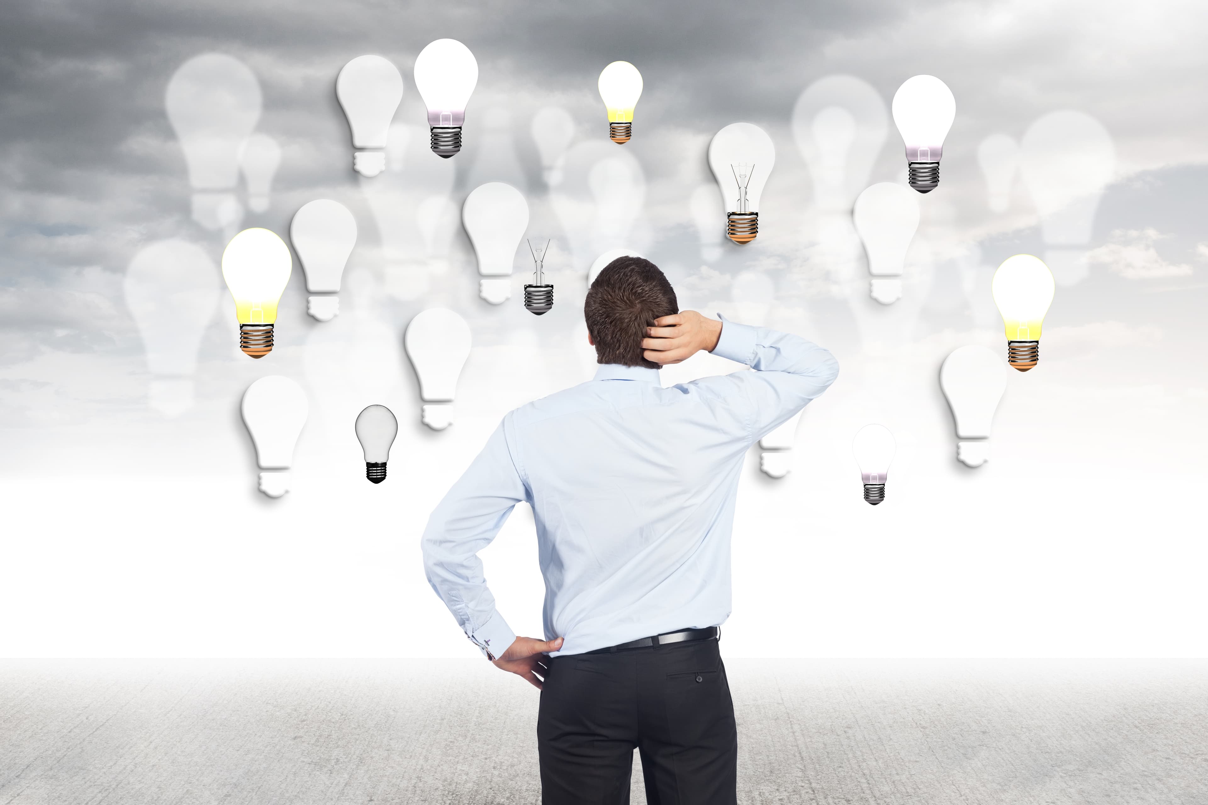 Um homem de pé de costas, com uma das mãos na cintura e a outra na cabeça, olhando para uma parede com luzes de bulbo, a procura de uma ideia brilhante.